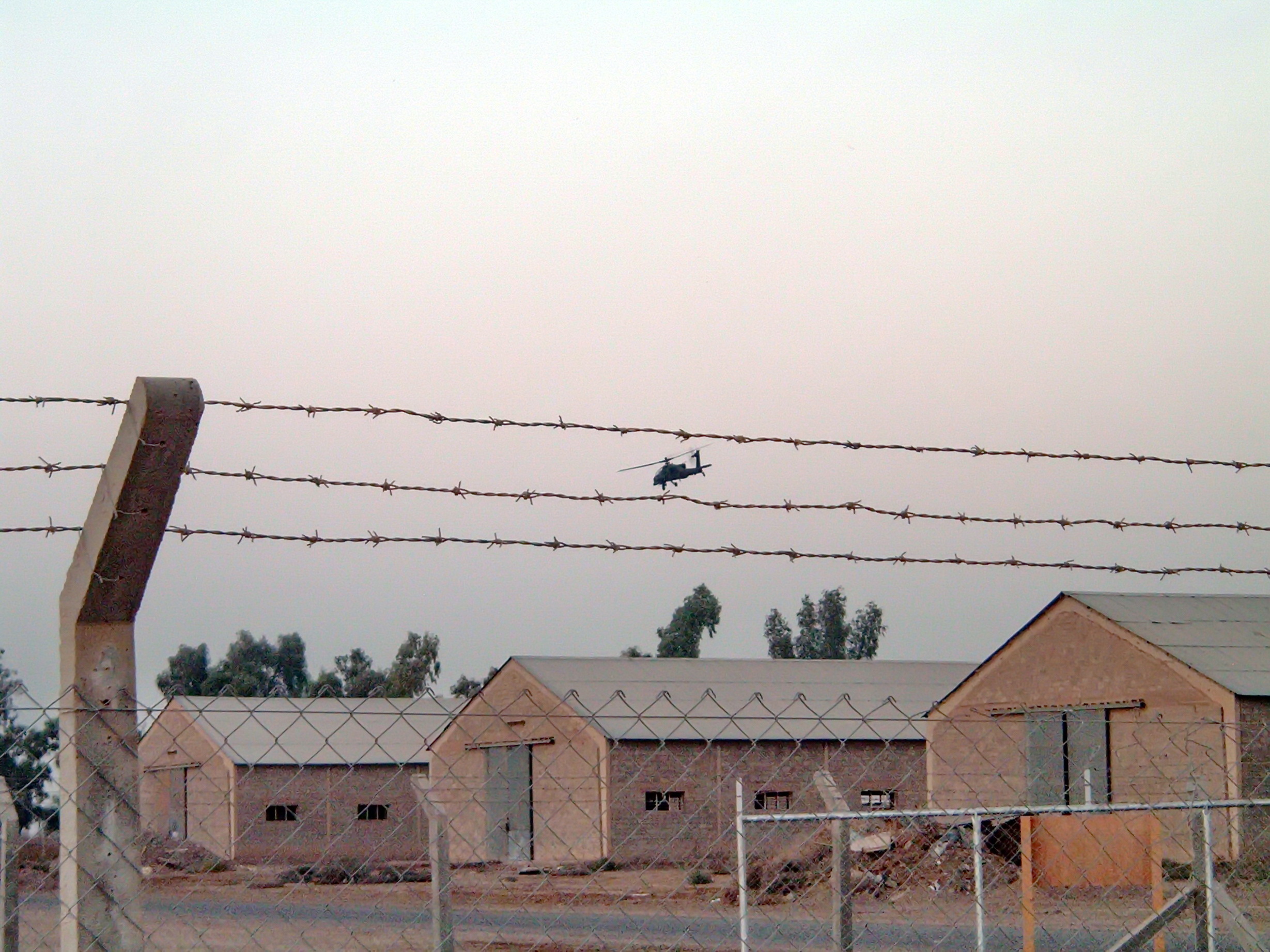 US Army helicopter flying above abandoned buildings at the end of a Forward Operating Base, framed by two strands of barbed wire