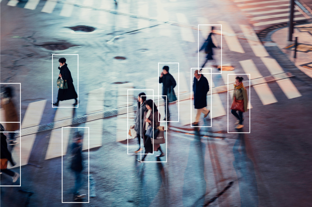 A nighttime street scene featuring people crossing a wet, reflective crosswalk. Each person is highlighted with a rectangular box, indicating some form of tracking or detection technology. The individuals are dressed in winter clothing, including coats and boots, and appear to be in motion, with some figures blurred to show movement. The background includes crosswalk lines and wet pavement, suggesting recent rain.