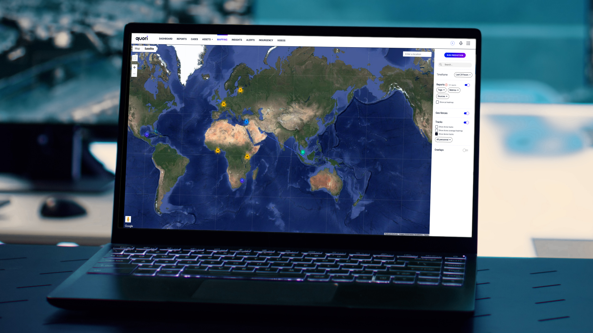 A laptop screen displays the Quori operational intelligence system web platform, featuring a detailed map of the world. The map shows different colored markers indicating activity or reports across various regions, including North America, Africa, Europe, and Asia. The user interface includes tabs such as "Dashboard," "Reports," "Cases," "Assets," "Mapping," "Insights," "Alerts," and "Insurgency" at the top, with additional filters and options for geofences and tracks on the right panel. The platform appears to offer real-time tracking and geospatial data.